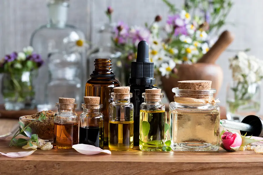 Assorted small glass bottles containing natural essential oils arranged on a wooden surface with flowers in the background