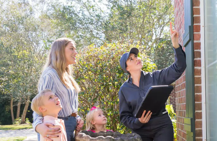 A pest control professional in a grey uniform points toward the upper corner of a brick house while a mother and two children look on.