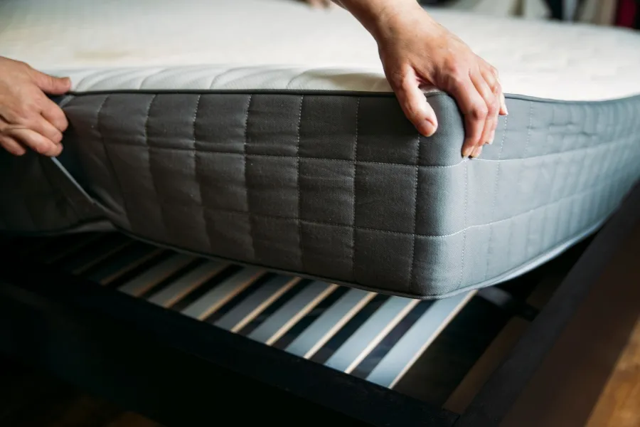 Hands lifting a mattress from a wooden slatted bed frame during a bed bug inspection