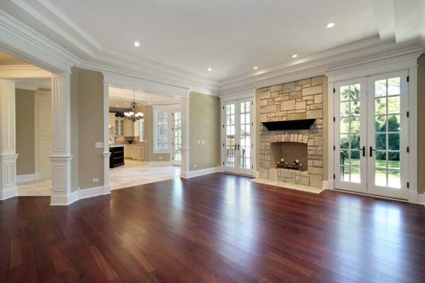 An empty living room with polished hardwood floors connects to a kitchen area. The room features a stone fireplace, large windows, and French doors leading to a grassy outdoor space.